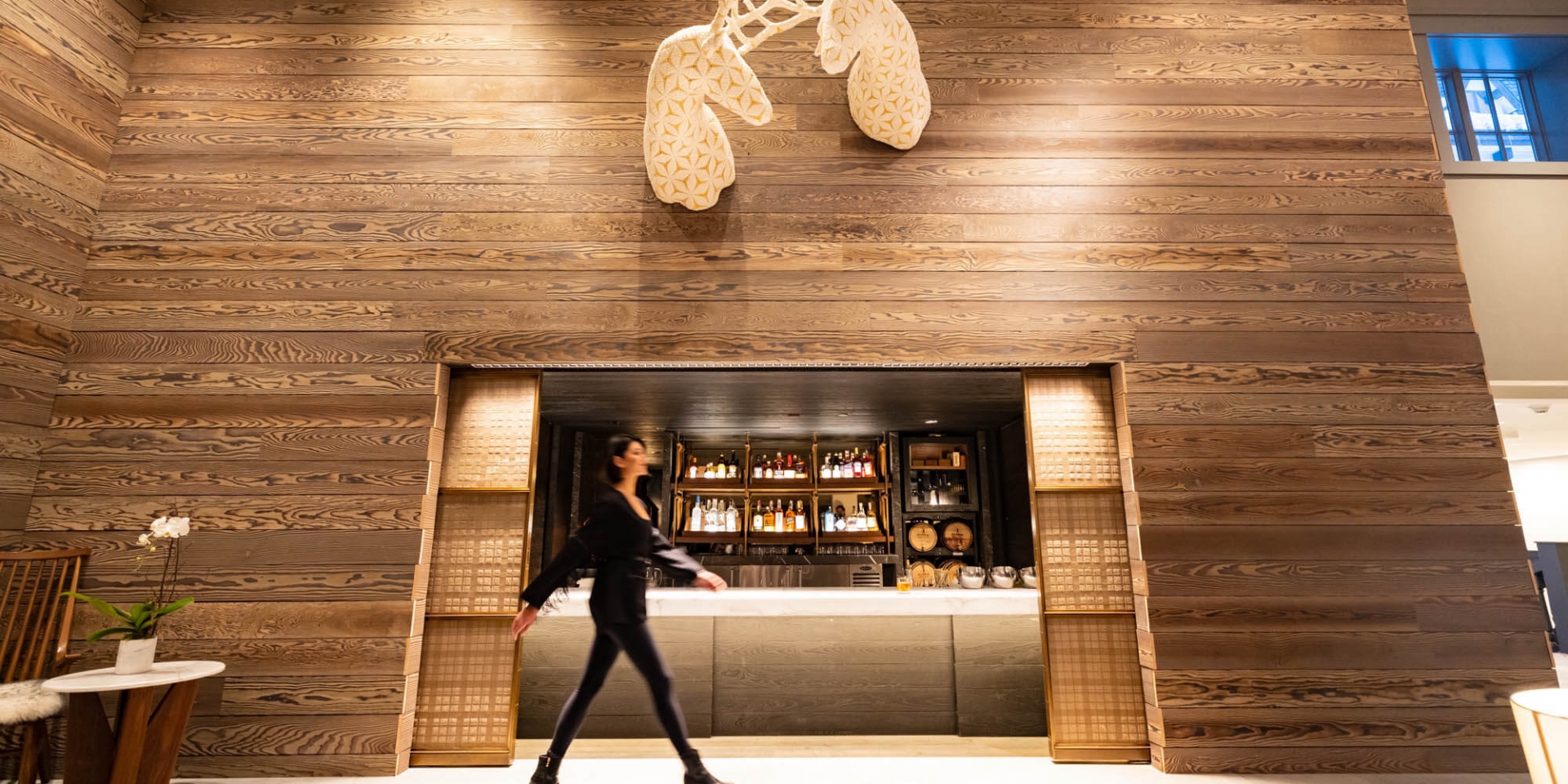 A person walks past a chic, wood-paneled lobby bar at The Hythe featuring a unique interlocking deer head sculpture.