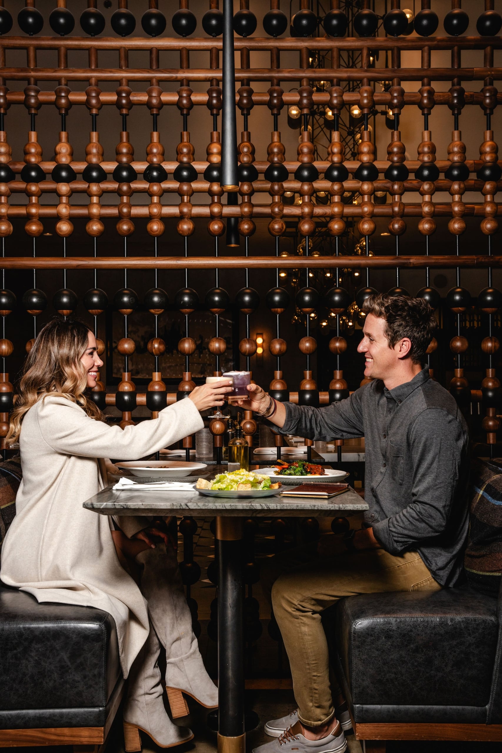 A couple toasts with cocktails at a marble table in front of a unique, dark wood architectural abacus-style wall.