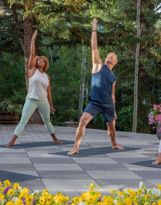 A group of four people practices yoga in warrior pose on an outdoor stone patio surrounded by lush summer greenery.
