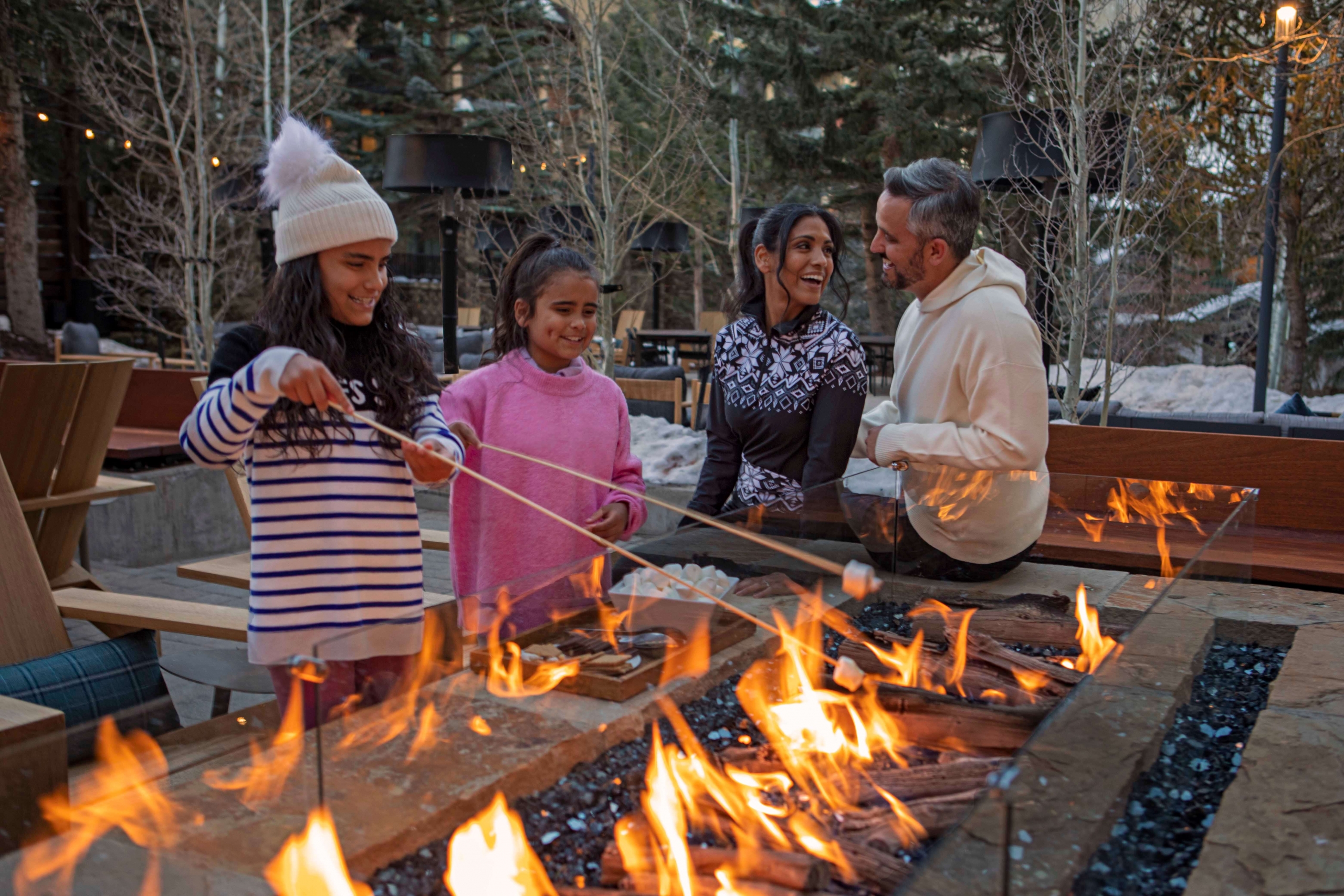 A happy family roasting marshmallows over a linear outdoor fire pit on a cozy, snowy evening at The Hythe.